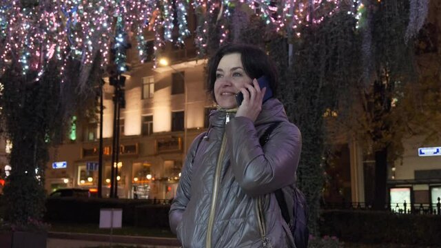 A middle-aged Caucasian woman in a lilac jacket takes out a smartphone and talks. New Year's illumination. in evening time