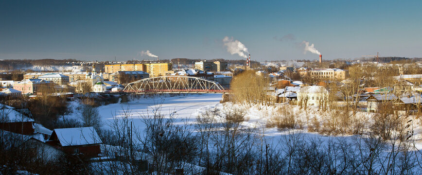 The cityscape of russian city Kungur of Perm Krai: a bridge over the frozen river, bare trees on its bank and many houses and smoky chimneys in the background in snowy winter.