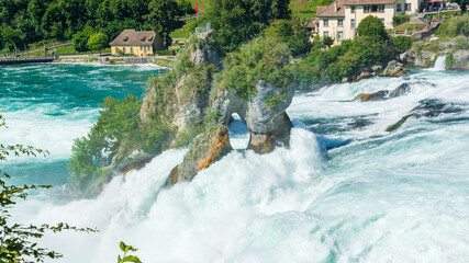 Panorama des cascades du Rhin ou Rheinfall en Suisse par une journée ensoleillée avec un ciel bleu avec les rochers et un village et les embruns du fleuve.