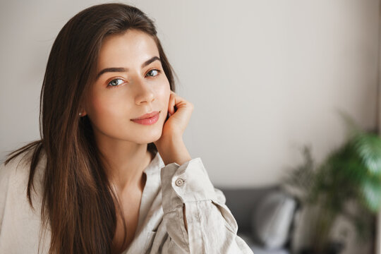 Young elegant woman smiling at camera, sitting at home with dreamy face. Girl with thoughtful face gazing at you