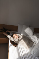 Vertical shot of beautiful woman waking up happy, stretching hands after good night sleep. Girl lying in bed and smiling
