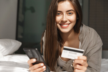 Close-up of smiling woman mobile shopping at home, lying in bed with credit card and smartphone