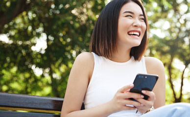 Happy asian girl sitting on bench and using mobile phone. Girl resting in park and browsing social...