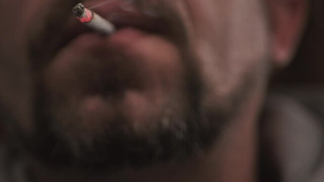 A man with a beard smokes a thin cigarette, the focus is on the face of the cigarette blurred