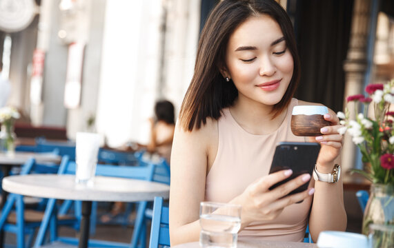 Charming asian woman with beautiful smile reading good news on mobile phone during rest in coffee shop, Happy female watching her photos on cell telephone while relaxing in cafe during free time