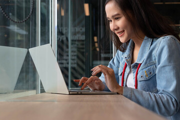 Young Asian woman wearing jeans jacket smiling. E - commerce business owner or freelance happy working use laptop Traveler blogger or remote work. Digital Nomad lifestyle.