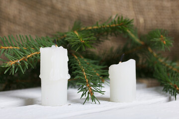 A candle in a glass candlestick. The candlestick is filled with pine cones and spices. Nearby are fir branches against the background of white pine boards.