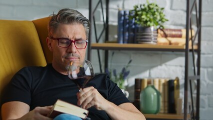 Older white man in glasses reading book at home sitting in armchair and drinking red wine in cosy room, smiling.