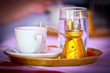 A closeup photo of a white cup, a glass of water and golden cezve on the tray on table.