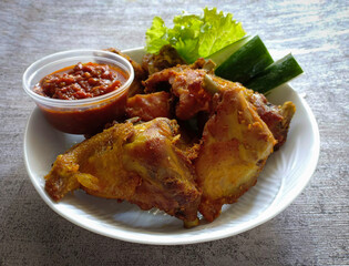 Indonesian food. Fried chicken served on a white plate along with pieces of cucumber, lettuce and a cup of cooked tomato chilli sauce. Very tasty, savory and delicious. 