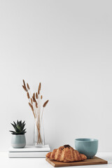 Croissant and a mug on a wooden stand, a vase and a pot with a plant on the books, white background. Eco-friendly materials in interior decor, minimalism. Copy space, mock up.