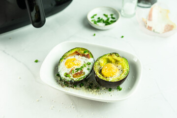 Baked eggs in avocado springkle with green onion and Parmesan cheese in a white dish, the Air Fryer and condiment behind, on the withe marble table. Healthy and keto breakfast. Eggs disk..