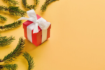 Gift box with a bow among the Christmas tree branches on a yellow background with copy space.