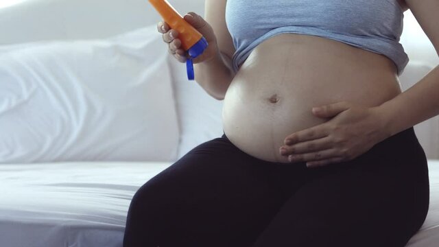 Young Asian pregnent mother holding lotion bottle and rubbing lotion around abdomen while sitting on bed
