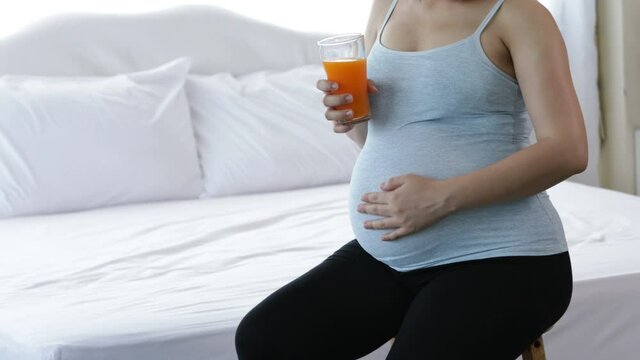 Young Asian pregnent mother holding orange juice and touch abdomen to talk with child inside while sitting on bed