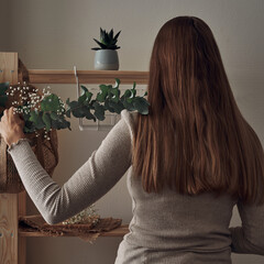 The girl decorates a wooden rack with an eco-bag with eucalyptus branches, flowers, houseplants. Eco-materials, storage. Folk.