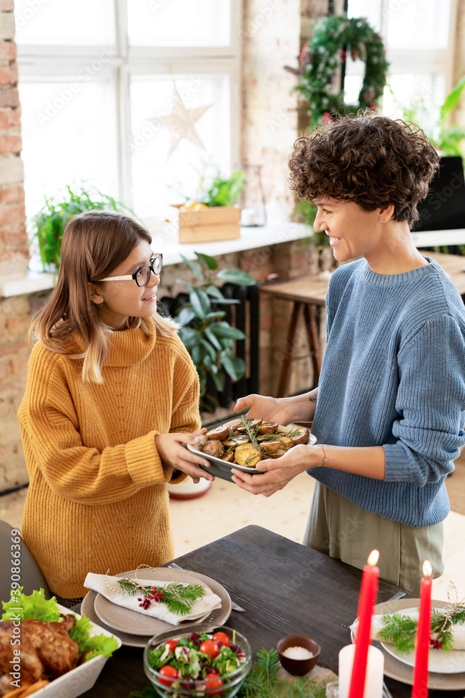 Wall mural happy young female passing her teenage daughter bowl with roasted potatoes