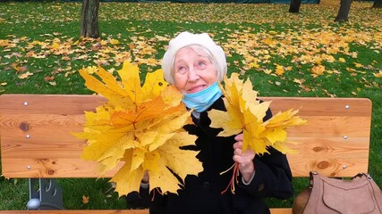 Older woman in protective mask and a bouquet of autumn leaves is having fun in the autumn park outdoors. COVID-2019 coronovirus. Health of the elderly. Portrait of 80 years old woman close up