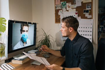 Fototapeta na wymiar Caucasian man wearing casual shirt using computer for telemedicine. Online doctors in video call. Telehealth during pandemic. Video consult in the computer monitor doctor wear medical face mask