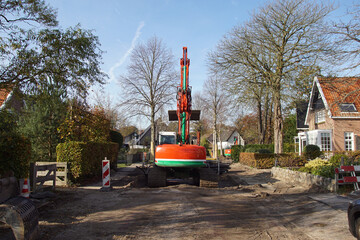 Fototapeta na wymiar Roadwork with a red, green excavator in the Dutch village of Bergen. Wood boards placed around the tree trunks and secured with wire to protect them from damage during the construction work. November,