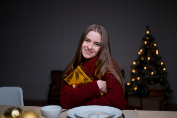 Caucasian girl sitting at a festive table with Christmas candles, hugging a gift box and rejoicing