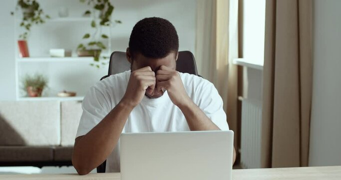 Portrait of serious upset tired black man African student guy looking at laptop in home office. Afro American freelancer feels stress reads bad news holds head with his hands in shock, loses job