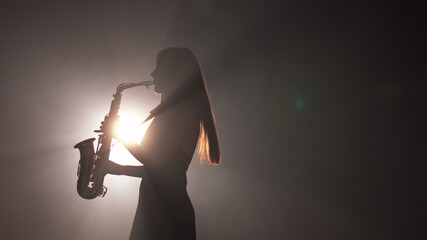 Young beautiful girl in a dark dress plays on a golden shiny saxophone on stage. Dark studio with...