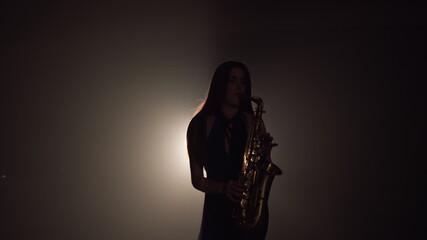 Young beautiful girl in a dark dress plays on a golden shiny saxophone on stage. Dark studio with smoke and stage lighting. Hands and saxophone close up. Side view.Slow motion video