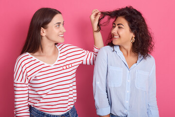 Girl pulling wavy hair of her friend, girls looking at each other with happy smile, females wearing casual attires, ladies wearing casual shirts posing isolated over pink background.