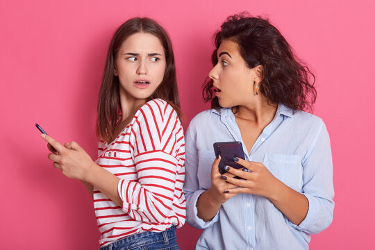 Shocked women reads stares at each other while holding cell phones in hands, female in striped shirt posing backwards to her friend, both girls being astonished, keeping mouth opened and has big eyes.