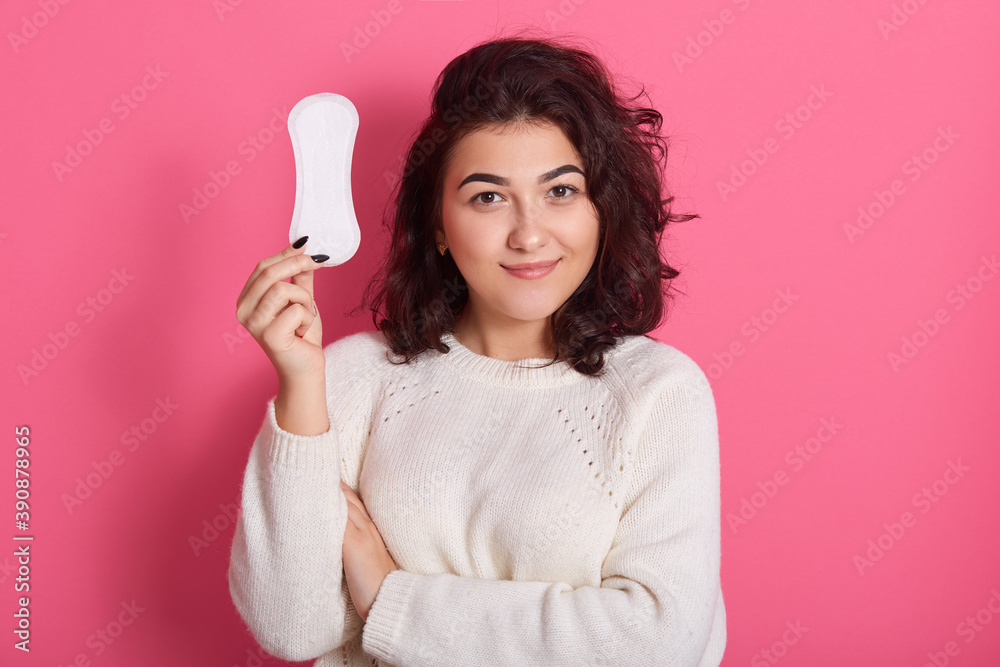 Wall mural Good looking feminine girl holds clean sanitary napkin, has menstrual cycle, happy to have good women health, female with dark wavy hair wearing white warm sweater isolated over pink background.