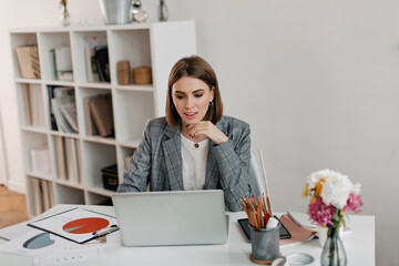 Close-up shot of adult woman in light-colored outfit looking enthusiastically at laptop. Business lady focused posing in white office