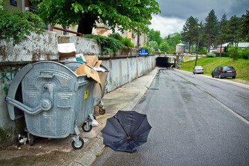 A road going under the tunnel surrounded with green trees with parked cars and a dumpster full of carton boxes and a black opened umbrella lying around in Cetinje, Montenegro.