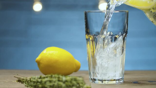 Close up pouring lemonade with thyme into a glass on blue background. Slide or move camera, dolly shot