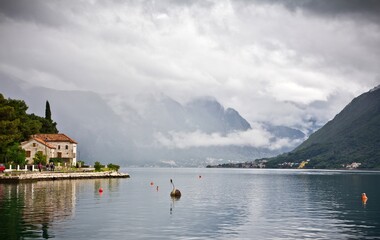 The stone embankment of the Bay of Kotor with buoys, small tile roof houses and mountains in clouds in the background in Perast, Montenegro.