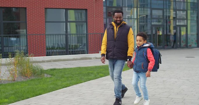 Happy little African American boy smiling and jumping while walking from school with father on street. Joyful male parent holding hand of his child. Dad going after lessons with cute kid with backpack
