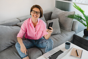 attractive smiling woman in pink shirt sitting relaxed on sofa at home at table working online on laptop from home freelancer social distancing self isolation communication holding phone