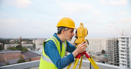 Caucasian senior man topographer in casque measuring angle with total station on roof of building. Male builder. Constructor doing topographic measures. Geodesy concept. Geodesic. Constructing work.