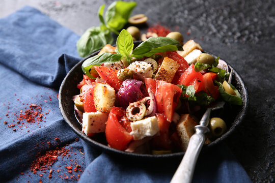 Oriental cuisine. Spicy vegetable salad with spices. Salad of tomatoes, olives, red onions, cheese and basil croutons on a black cast-iron plate on a black background with red spices. Copy space