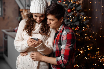 Happy young couple checking Christmas messages on a smartphone hugging standing near decorated christmas tree enjoying spending time together. Joyful cozy moments in winter holidays. Advertising
