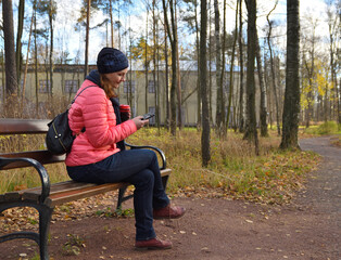 A woman sits on a bench in an autumn park, drinks hot coffee and looks into the smartphone screen