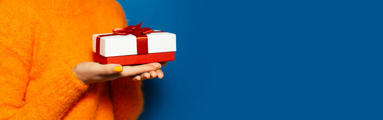 Close-up of female hands holding white gift box with red bow on background of blue color with copy space. Wearing orange sweater. Christmas concept.