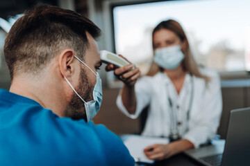 Fototapeta premium Beautiful female doctor wearing protective face mask, using infrared forehead thermometer gun to check patient's body temperature. Pandemic, Covid-19 concept.