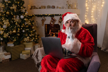 Modern Santa Claus. Cheerful Santa Claus working on laptop and puts a finger to his lips Shhh at his chair with fireplace and Christmas Tree in the background.
