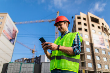 Portrait of young engineer man use phone while standing at construction area. Builder in helmet control according to plan by mobile phone.