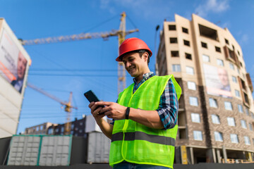 Fototapeta na wymiar Portrait of young engineer man use phone while standing at construction area. Builder in helmet control according to plan by mobile phone.