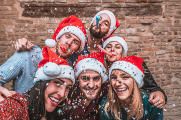 Group of young friends wearing santa hats having fun at party, posing for photos - Happy young people blowing party whistle at a new year's eve party -  Confetti falling in the air