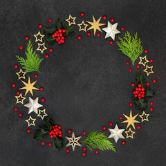 Christmas wreath decoration with winter greenery of holly & loose berries, cedar cypress fir & gold star decorations on grey grunge background. Abstract composition for the xmas & solstice season.