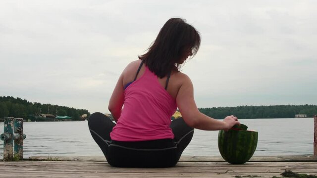 Back view of woman in pibk tank sitting on old pier with watermelon behind her. She takes a slice of watermelon and eats it. Summer food.