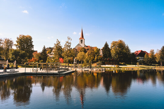 Cityscape view of the village of Schonach in the Black Forest of Germany during autumn. A church is in the centre and reflects in the water of a lake.
